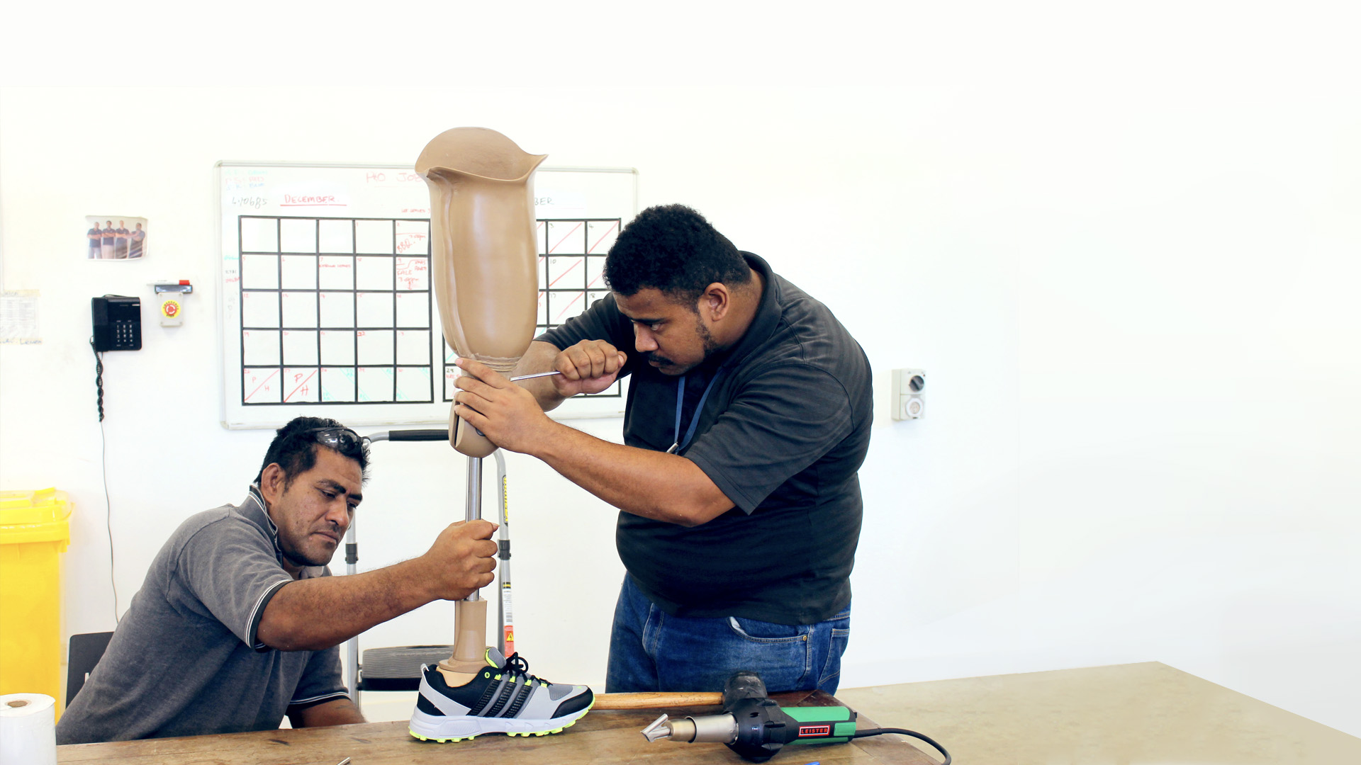 Two men work together to make an adjustment to a prosthetic leg placed upon a workbench. One man sits holding the leg while the other stands, working on it with a tool. Both look very focused.