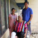 A woman using a wheelchair with a bandage around one leg smiles for a photo with two service providers who stand next to her.