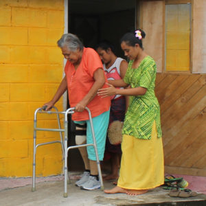 A woman learning to use her prosthetic leg walking outside her house, supported by one of the MDS staff members.