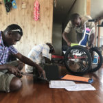 Three personnel work on a wheelchair in a clients home. One is measuring a cushion to cut, two are checking the nuts and bolts!