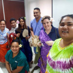 A group selfie shows 5 women and two men smiling at the camera. The nurses are wearing scrubs. Nalini and Katrina are holding gifts including lavalavas, fans, and flowers.