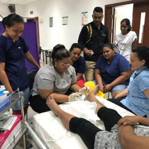 DFC nurse Tai is debriding the foot wound of a client whose face we can’t see. To her right is a dressings trolley. She is being observed by five nurses and Podiatrist Nalini.