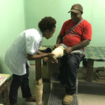 A man in a red t-shirt and cap is sitting on a treatment table; he has a right below knee amputation. A lady in a white coat is fitting a liner to his stump, next to him we can see a prosthetic device ready to be fitted, on the floor.