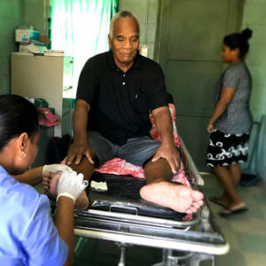 A photo of diabetic foot care in action! A Kiribati man is sitting on a clinic bed, while a female nurse, wearing a blue nurses uniform and disposable gloves treats his foot.
