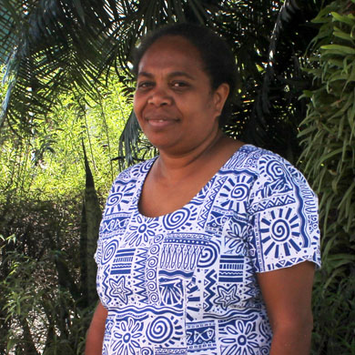 Profile photo of Elsie Taloafiri from the Solomon Islands, wearing a blue and white patterned dress. She is standing in front of tropical trees.