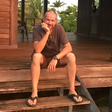 Ray Mines sits on the steps of his wooden home. The sun is setting, giving the photo a glow. In the background are tropical trees including coconut trees.