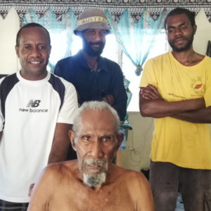 Three ni-Vanuatu men stand in the background. Another ni-vanuatu man is sitting in front of them in a wheelchair, but you cannot see the wheelchair in the photo. One of the men is leaning on the push-handle of the wheelchair.