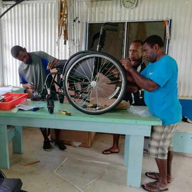 Three Ni-Vanuatu men are assembling a wheelchair, on top of a blue, wooden table.