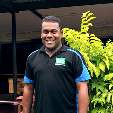 Jope stands outside of Frank Hilton Organisation smiling at the camera. He is wearing a black and blue uniform work shirt and a traditional Fijian mens sulu vakatoga. There is a lush green garden in the background.