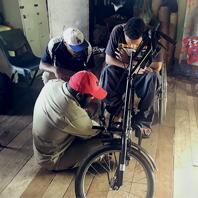 Samuel, sitting in his wheelchair with a tricycle attachment. Sammy and Alfred are adjusting his footplates to improve the angle.