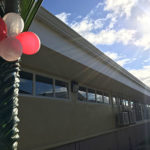 A photo of the outside of the building, with a decorated post in the foreground. It is wrapped with palm fronds and tied with balloons for the opening ceremony.