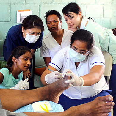 A client from Kiribati is having their foot wound treated by a Kiribati nurse during a training clinic. She is being observed by three of her team, and one MA volunteer. They are all in a small, huddled group around the end of the treatment bed.