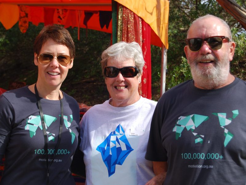 Three people wearing sunglasses stand side by side, smiling. Two are wearing an MA shirt with a green world map. One is wearing a MA shirt with a blue iceberg.