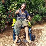 A man using a new wheelchair sits outside in the shade of green plants, he smiles widely and gives a big thumbs up to the camera.