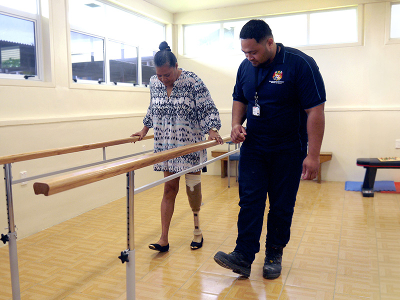 A woman using a prosthetic uses parallel bars to support herself as she walks. A service provider walks beside her. They both have the same foot out.