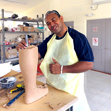 Vai'uli stands at a workbench in an apron, holding a unfinished lower-limb prosthetic. He is smiling brightly and giving a thumbs up.