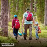 A man and two children walk through a forest. The man holds a child's hand. They are all wearing backpacks.