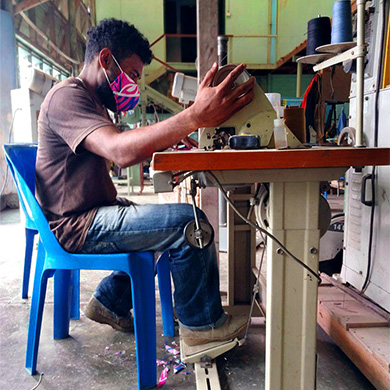 A man sits at a sewing machine. He is wearing a colourful face mask and looks very focused on the task.