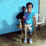 Violet stands in colourful shirt and shorts, using crutches to help her stand with her first prosthesis. Her mother sits behind her. Both are smiling brightly.