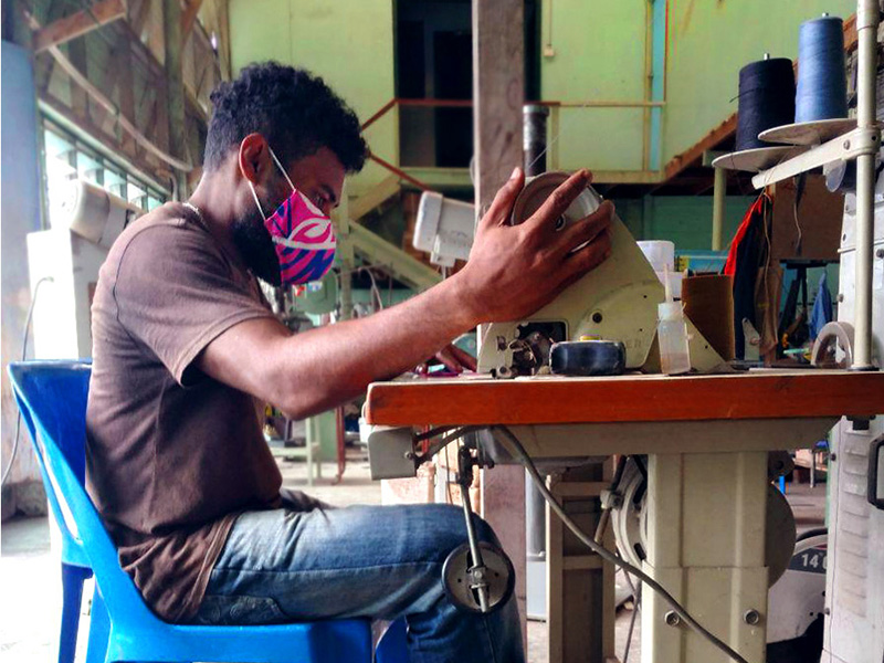 A man sits at a sewing machine. He is wearing a colourful face mask and looks very focused on the task.