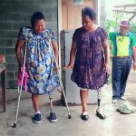 Two women, both with lower limb amputations and using a prosthesis, stand side by side. One woman is using crutches and looking down at her feet, while the other woman stands unsupported and is speaking encouragingly.