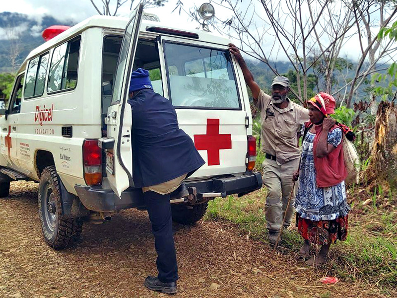 On the side of a dirt road, surrounded by forest and mountains, a large car with a red cross on it has stopped. A man looks in the back of it, while a woman using a thin stick as a cane, and another man wait patiently.