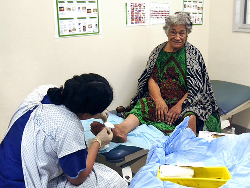An older woman sits up on a treatment bed while a woman dressed in uniform, protective apron and gloves treats a wound on her foot. Next to them is a colourful set of equipment to treat a wound.