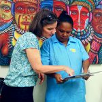 A woman in a nurse uniform holds a clipboard, another woman standing next to her points at it. They are both looking at the clipboard carefully. They stand in front of a colourful painting of people in traditional Papua New Guinea dress.