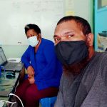 A man and a woman take a selfie. They are wearing facemasks and sat at a desk, socially distanced to work.