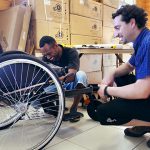 Two men work together to build a wheelchair. One man leans down to adjust something with a smile, the other watches closely, also smiling.
