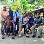 9 people, some sitting and some standing, are outside in the shade smiling at the camera. One man is using a wheelchair and a woman in the centre is using a lower-limb prosthesis.