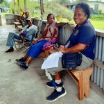 Almah, in NOPS uniform and using a lower limb prosthesis, sits on a bench under a verandah with three other people who are all using walking aids and/or prosthetic lower limbs. All are smiling for the camera.