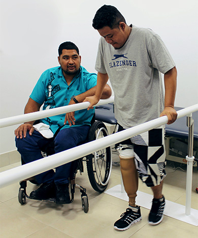 A man in a bright shirt uses a wheelchair and sits alongside a man using parallell bars and a lower limb prosthesis to walk. The man in the bright shirt is speaking encouragingly.