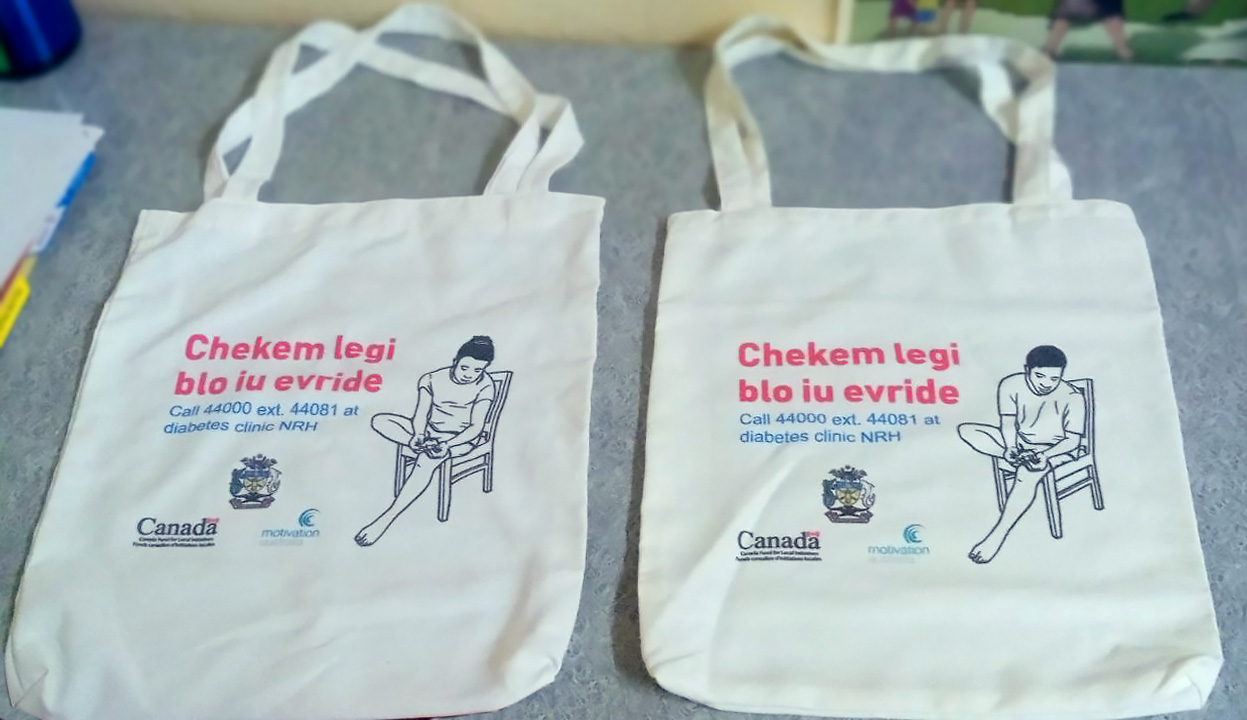 Two white bags with a graphic print. The graphic shows a man or women checking their feet, the phrase "Checkem legi blo iu evride'the number of the diabetes clinic, and logos for the Ministry of Health, CFLI and Motivation Australia.