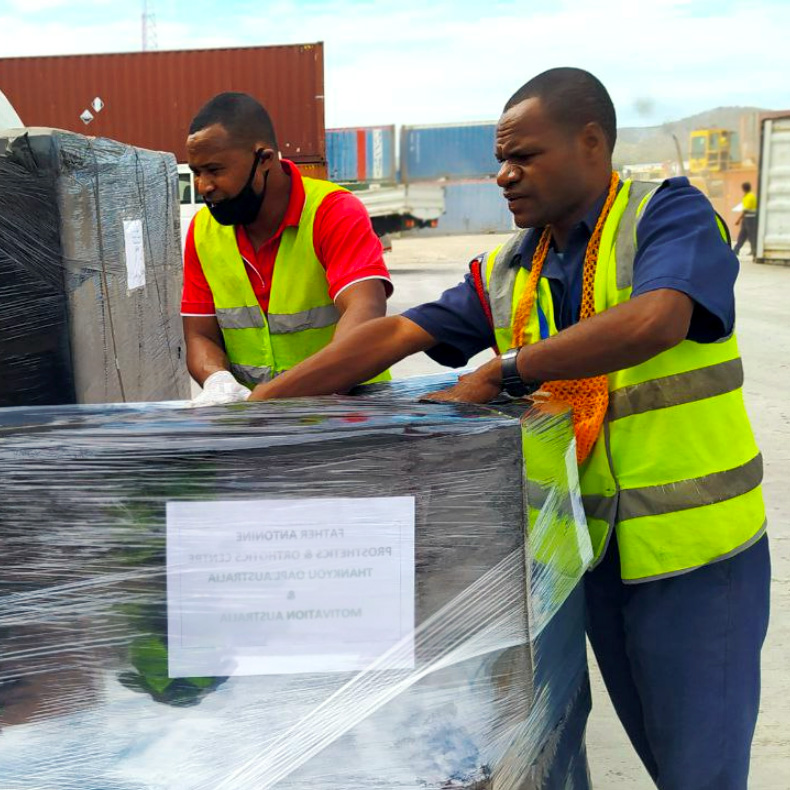 Two men in bright vests push a large wrapped package in a shipyard.