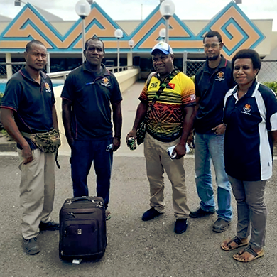 A group of five people stand outside. Four are wearing the same uniform, and one man is wearing a brightly coloured shirt. This man has a suitcase and backpack with him.