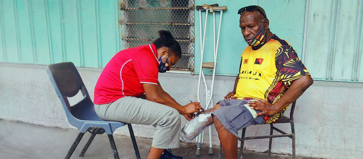 A man sits opposite a health worker who is wrapping his residual lower limb in plastic. A pair of crutches are propped against the wall nearby.