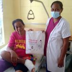 A woman with her foot bandaged sits on a hospital bed, holding up a foot care kit tote bag with a health worker.