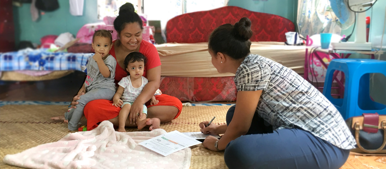 A woman sits on the floor, holding two small children in her lap. She smiles at a health worker who sits nearby, writing in forms as the other woman speaks.